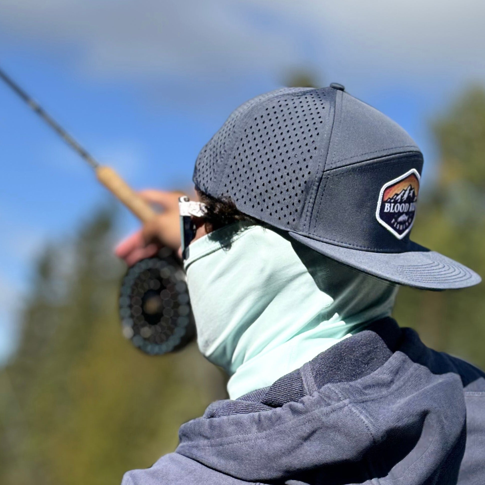 Caucasian 23 year old male centerpin float fishing for salmon on a river in Michigan wearing a grey blue men's snapback fishing trucker hat with Blood Run Fishing Sun Mountain logo patch on the crown, wearing a light green UPF50 fishing neck gaitor and blue grey french terry men's midweight cotton hoodie with Blood Run Fishing logo on the back wearing Pit Viper sunglasses.
